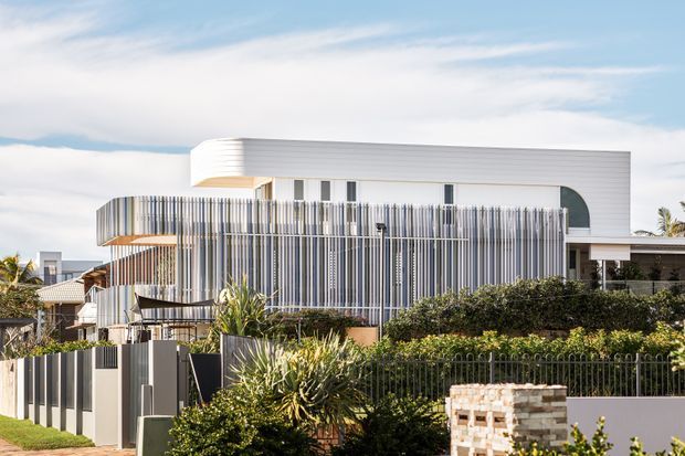 Contemporary multi-level residential facade with a rounded white upper volume, extensive vertical screening, projecting balconies, and layered landscaped foreground. The architecture combines clean horizontal lines with curved corners and a highly articulated striped facade treatment.