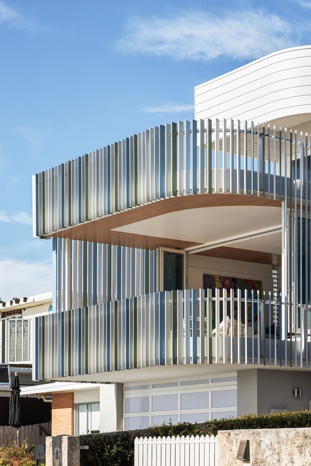 Exterior view of a contemporary multi-level residence with prominent curved balcony edges, extensive vertical metal privacy fins, white rendered walls, timber-lined soffits, and large glazed openings. The building sits in an urban residential setting under a bright blue sky, with a garage and front boundary landscaping visible at ground level.