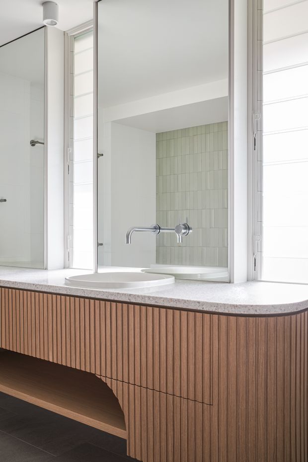 Bright contemporary bathroom vanity with a large mirrored wall, white vessel basin, wall-mounted chrome tapware, and a pale green vertically tiled niche visible in reflection. The custom vanity has a curved white stone benchtop and fluted mid-tone timber cabinetry with an open recessed shelf. White plantation shutters and minimal cylindrical ceiling lighting complete the clean, refined fit-out.