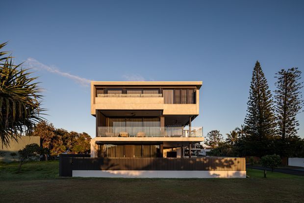 A contemporary multi-storey residential house viewed from the front across a lawn. The building has a strong rectilinear form with projecting upper levels, extensive dark-framed glazing, recessed balconies, vertical timber screening, and warm-toned cladding. A dark perimeter fence, mature palms, and tall conifers frame the property under a clear blue sky.