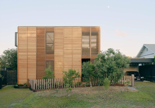 Front exterior view of a contemporary two-storey residential building clad in warm horizontal timber boards, with tall louvred timber screens over the windows and a recessed dark entry or opening. The house sits behind a low timber picket fence and landscaped planting, with a separate carport and neighbouring gabled structure visible to the right. The composition is clean and modern, photographed in soft daylight.