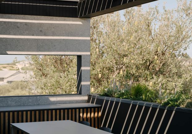 A contemporary covered outdoor dining terrace or balcony with dark metal cladding, concrete walls, timber-look vertical screening, and a large window opening. A dining table occupies the foreground, while dense planting and a residential landscape are visible beyond the balustrade. Strong sunlight creates diagonal shadows across the dark surfaces.