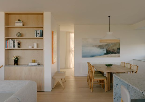Bright open-plan interior showing a dining area beside a kitchen island, with pale timber flooring, built-in timber shelving and cabinetry, a light neutral palette, and coastal artwork. A partial living area and hallway are also visible.