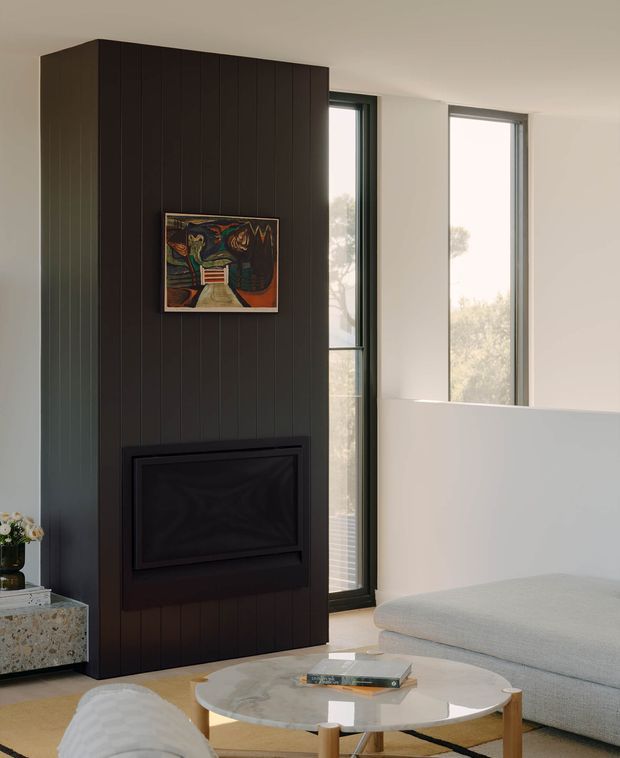 A refined living room centred on a tall dark vertical-panel feature wall with an integrated black fireplace and artwork. Narrow floor-to-ceiling windows flank the feature wall, while a pale curved sectional sofa, round marble-topped coffee table, and terrazzo side table create a soft, contemporary interior.