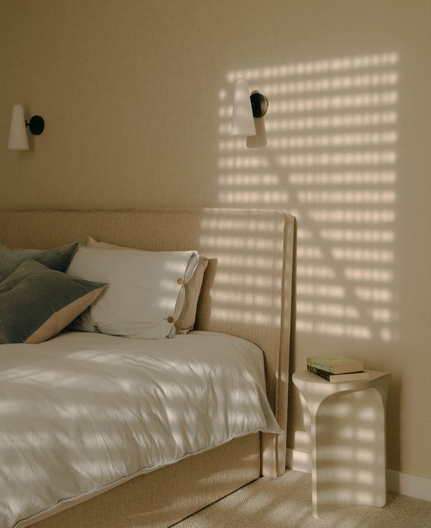 A softly lit, warm-toned bedroom featuring a large upholstered bed with a tall textured beige headboard, white bedding, and muted green and beige pillows. Minimal white wall sconces flank the bed, while patterned sunlight from blinds creates a dramatic grid of light and shadow across the wall, bed, and curved bedside table. The room has a calm, refined, contemporary feel.