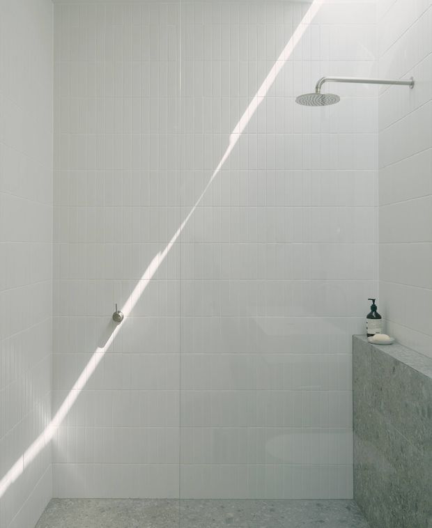Minimalist walk-in shower with white rectangular wall tiles, a brushed metal rainfall showerhead and controls, and a grey terrazzo or stone ledge. A strong diagonal beam of sunlight crosses the otherwise bright, restrained space.