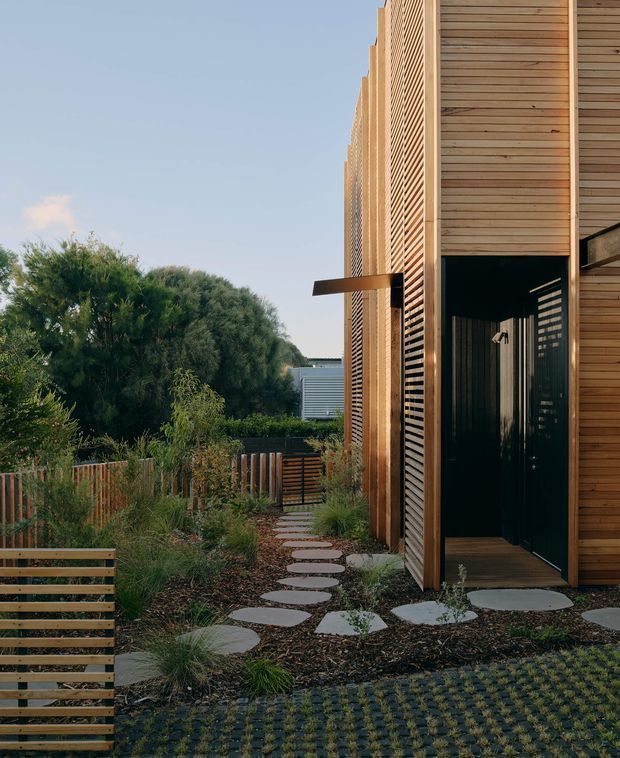 Exterior view of a contemporary timber-clad building entrance set within a landscaped garden. The facade uses vertical and horizontal timber slats, with a dark recessed doorway and timber decking. A stepping-stone path, native-style planting, timber fencing, and permeable patterned paving lead toward the entrance.