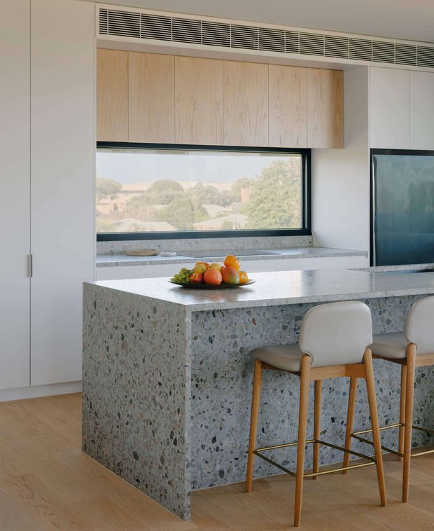 Bright contemporary kitchen featuring a large island clad in grey terrazzo, pale timber cabinetry, integrated ventilation, light timber flooring, and a wide black-framed window overlooking a leafy suburban setting. Two upholstered timber bar stools sit at the island, which has a terrazzo benchtop and waterfall sides.