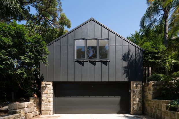 Exterior view of a compact gable-roofed building, likely a garage or studio, clad in dark vertical panels. A broad dark garage door occupies the lower level, with a small horizontal window above, flanked by rough pale stone masonry walls and surrounded by mature greenery and palm trees.