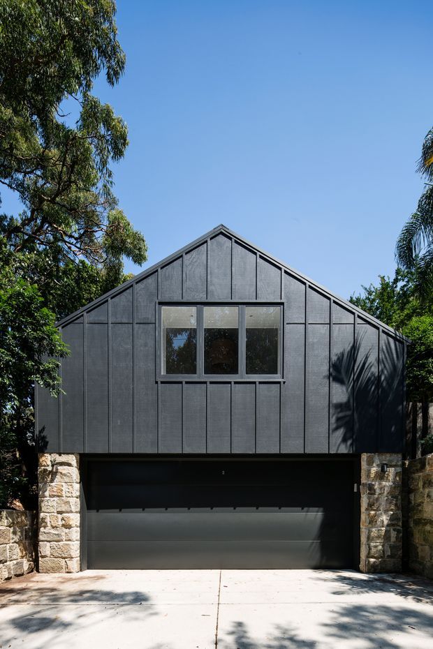 Front exterior view of a compact gable-roofed garage or secondary structure. The facade is clad in dark charcoal vertical panels with a centered row of windows, a wide dark garage door at ground level, and pale rough-cut stone side walls. Mature trees frame the building and cast shadows across the driveway.