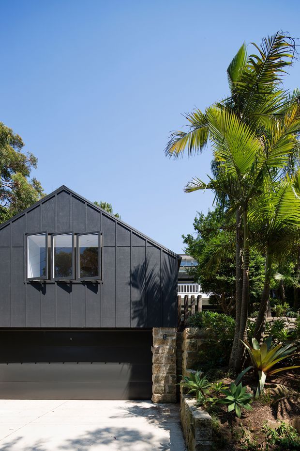 Exterior view of a compact gable-roofed home with dark vertical cladding, projecting rectangular windows, and a large dark garage door. A stone retaining wall, concrete driveway, and lush tropical planting with palms frame the facade in bright sunlight.