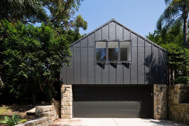 Front exterior view of a compact gable-roofed building integrated into a landscaped site. The upper facade is clad in dark vertical panels with a projecting multi-pane window, while the lower level contains a wide dark garage door framed by rough natural-stone masonry piers and retaining walls. Dense greenery surrounds the structure.