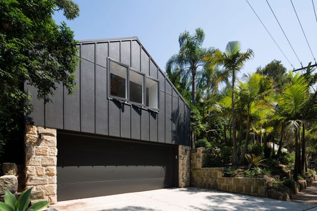 Exterior view of a contemporary elevated residential garage and upper-level facade. The building features dark vertical cladding, a projecting awning-style window, substantial light stone masonry piers and retaining walls, and a wide dark garage door. Lush tropical landscaping surrounds the driveway, with overhead utility lines visible.