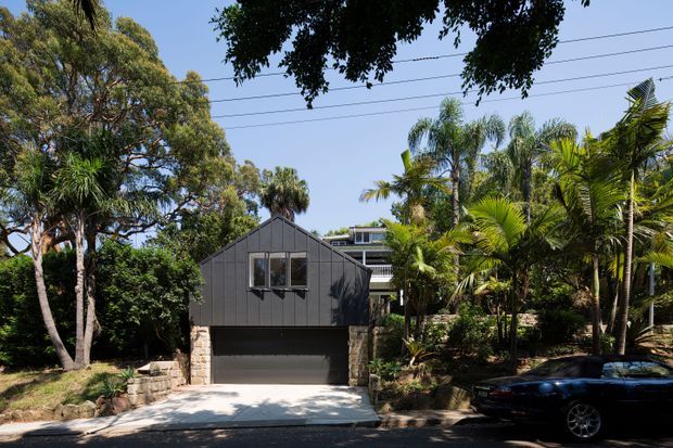 Street-facing exterior of a contemporary dark-clad gabled garage or outbuilding, with a wide integrated garage door, stone retaining walls, lush tropical landscaping, and a larger residence partially visible behind it. The property is photographed from the road in bright daylight, with overhead utility lines and a parked car in the foreground.