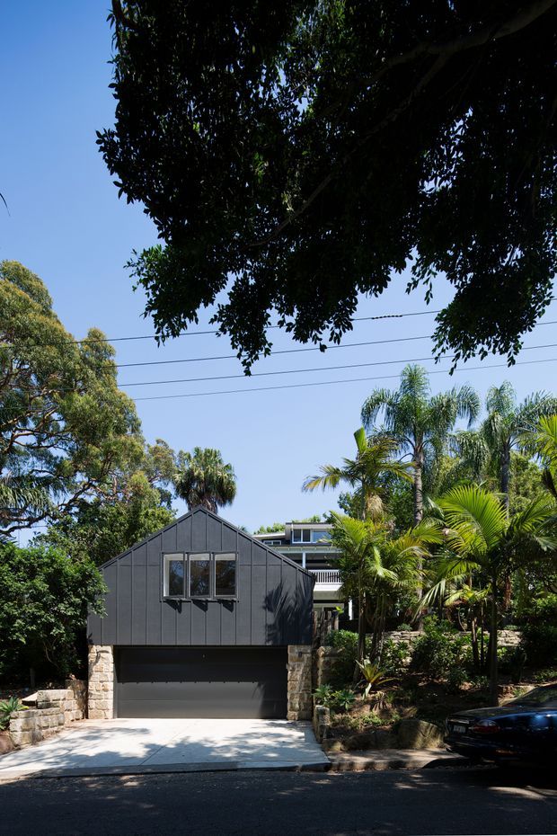 Street-facing exterior view of a compact dark-clad garage or outbuilding with a steep gable roof, integrated wide garage door, and a small upper window. Rough light stone pillars and retaining walls frame the structure, while dense tropical and subtropical landscaping surrounds a larger residential building partially visible behind it. Overhead utility wires and a paved driveway are visible.