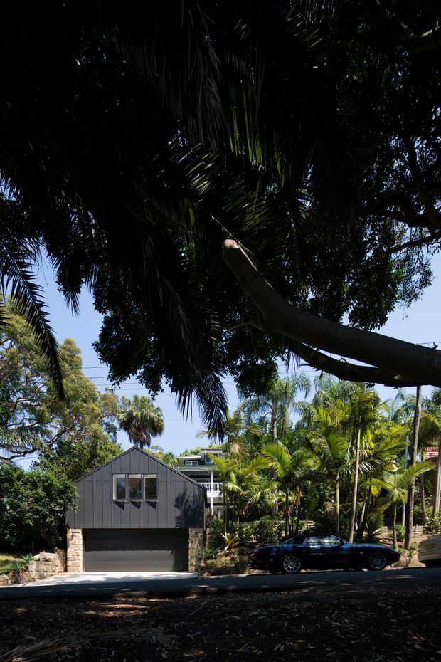 Street-facing view of a compact modern dark-clad residential garage or outbuilding with a steep gabled roof, upper window, and wide garage door set on a light stone base. The building is surrounded by dense tropical landscaping and mature palm trees, with parked cars along the street in the foreground.