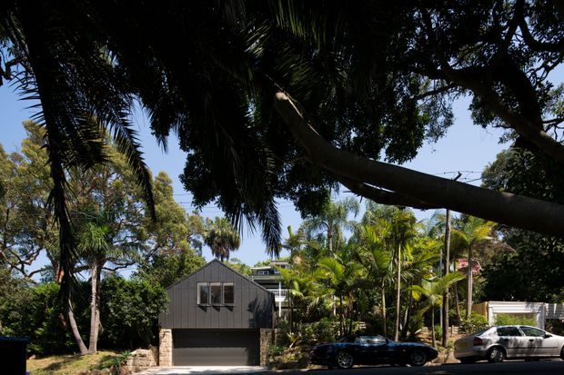 Street-level exterior view of a compact dark-clad residential house partially screened by mature tropical and subtropical planting. The home has a simple steep gabled form, a large garage opening at ground level, upper-level glazing, and a light stone retaining wall. Parked cars and dense trees occupy the foreground and surrounding streetscape.