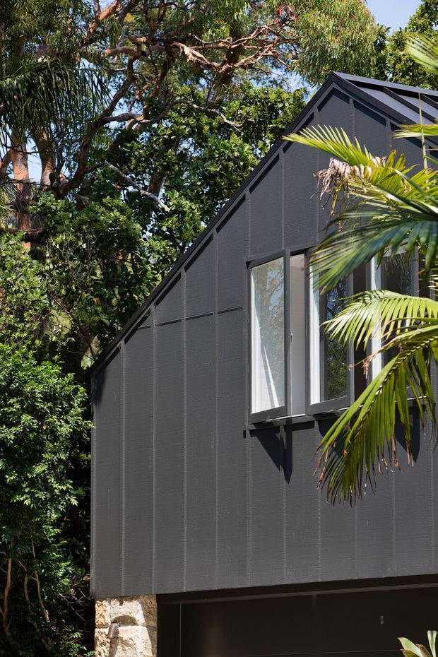 Exterior detail of a dark, steeply gabled building nestled among dense tropical vegetation. The facade has fine-textured dark cladding, sharply expressed gable trim, tall projecting windows with dark frames, and a stone base visible at ground level. Bright palm fronds and mature trees surround the structure.