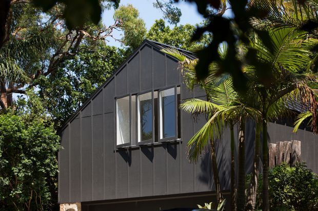 Exterior view of a compact, elevated dark-clad building set among dense tropical and subtropical vegetation. The facade has a steep gable roof, vertically proportioned dark panels, and a projecting bank of tall windows with simple dark frames. Stone or masonry supports are partially visible at the base. The image focuses on the building envelope and roofline, with landscaping framing the structure.