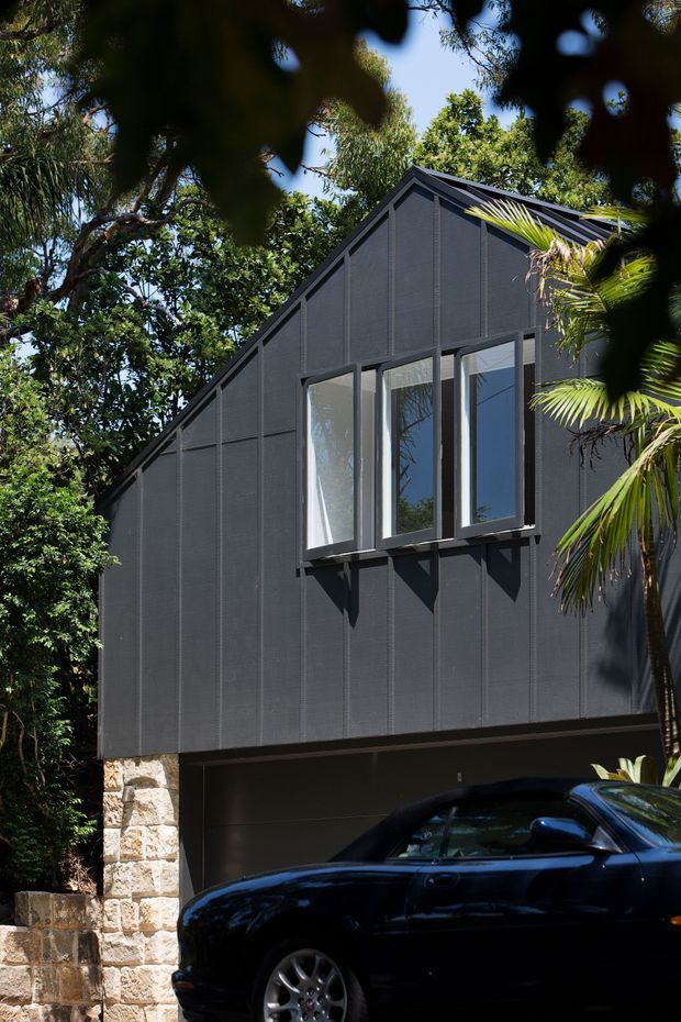 Exterior architectural photograph of a compact, dark-clad building set among lush tropical vegetation. The facade has a steep gabled roof, vertically detailed dark panels, tall narrow windows with projecting frames, and a stone-clad masonry base. A shaded garage or carport opening is visible at ground level, partly obscured by a parked car.