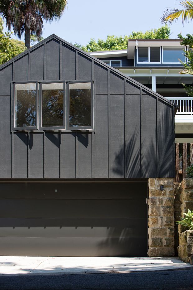 Exterior view of a contemporary residential addition or garage with a steep gable form and dark textured cladding. Three projecting windows punctuate the facade above a dark garage door, while a lighter multi-level house with balcony railings is visible behind. A rough natural-stone retaining wall frames the driveway, with tropical vegetation around the property.