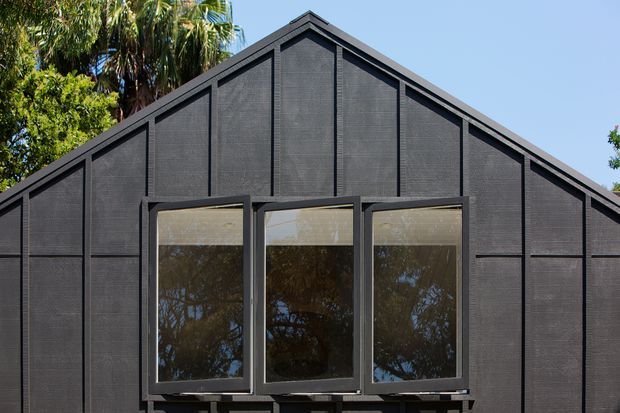 Close-up exterior view of a dark, gable-ended building facade with vertically framed projecting windows. The facade has a textured charcoal-black finish with prominent dark framing and a simple pitched roofline. Reflections of trees are visible in the glazing, with palm foliage surrounding the structure.