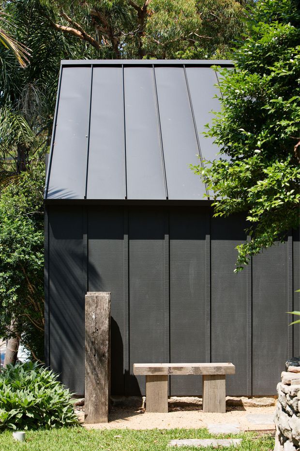 A compact dark-clad garden structure or outbuilding set among dense tropical and native vegetation. The facade uses vertical dark panels with a large light grey standing-seam metal pitched roof. A simple timber bench and weathered timber post sit in front, with a low rough-stone wall at the side and a small gravel entry area. The image is photographed outdoors in bright natural light.