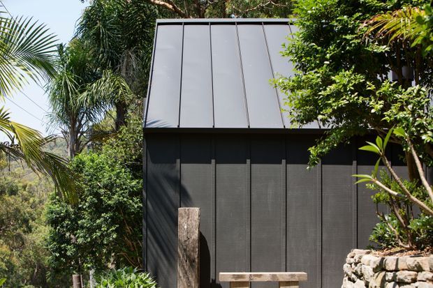 Exterior view of a compact contemporary structure set within lush tropical landscaping. The dark vertically panelled facade has a large steeply sloped metal roof or upper wall plane, with a low stone retaining wall and timber elements in the foreground.