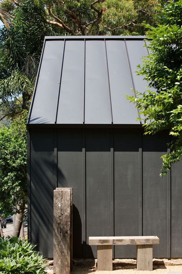 Exterior view of a compact dark-clad structure set within dense greenery. The facade uses vertically panelled dark grey cladding with prominent black vertical seams, topped by a steeply sloped pale grey metal roof or upper facade plane. A simple timber bench and rough timber post sit in front, with a suburban street partially visible at the edge.