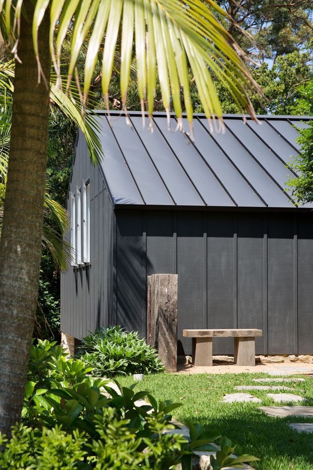 Exterior view of a compact contemporary garden building surrounded by dense tropical landscaping. The structure has dark vertical cladding, a light grey standing-seam metal pitched roof, narrow vertical windows, and a simple timber-and-stone bench beside a stepping-stone path. Palm foliage frames the foreground.