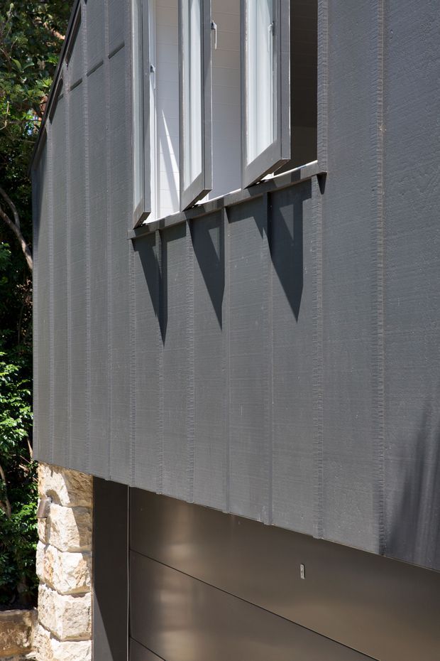 Close-up exterior view of a contemporary building facade. The wall is clad in dark grey vertically textured panels, with projecting white-framed awning windows casting strong geometric shadows. A section of rough natural stone masonry and a dark flush panelled lower wall are visible at the base, with dense greenery at the side.