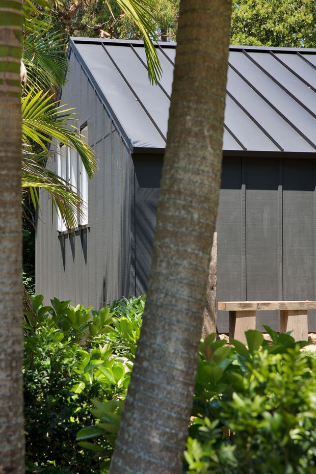 A close exterior view of a contemporary dark-clad building nestled among lush tropical planting and palm trees. The structure has a sharply pitched metal roof with prominent standing seams, dark vertical wall cladding, screened or louvred side panels, and a small timber railing or deck edge in the foreground. The composition is framed by palm trunks and dense greenery.