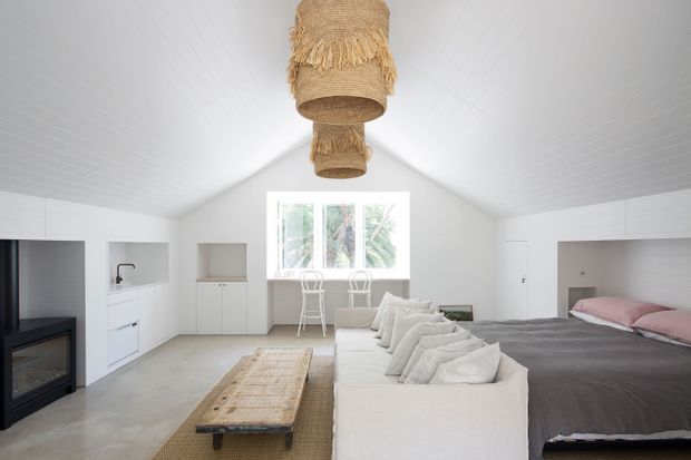 Bright open-plan studio-style interior with a white pitched ceiling and walls, pale concrete flooring, integrated white cabinetry, a compact kitchenette, a lounge area with a white sofa, and a bed positioned at the right. A large central window overlooks greenery, while woven pendant lights, a timber coffee table, and neutral textiles add natural texture.