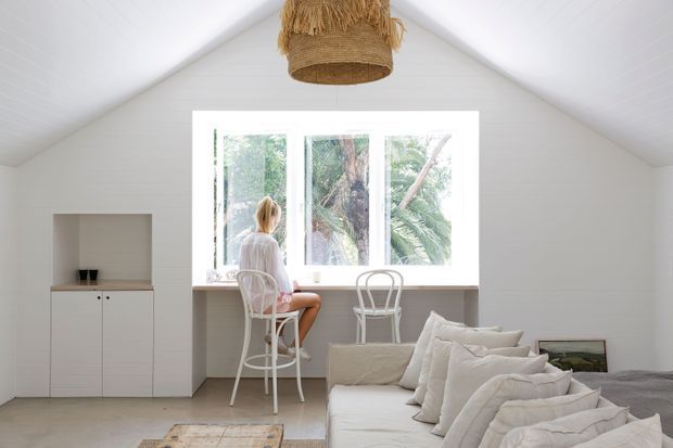 Bright, airy attic-style living space with a steeply pitched white paneled ceiling and matching walls. A large bank of windows overlooks lush greenery, with a narrow built-in counter and two white stools creating a casual workspace or breakfast perch. White built-in cabinetry sits to the left, while a white upholstered sofa with layered neutral cushions occupies the foreground. A woven pendant light adds natural texture, and the overall styling is relaxed and coastal.