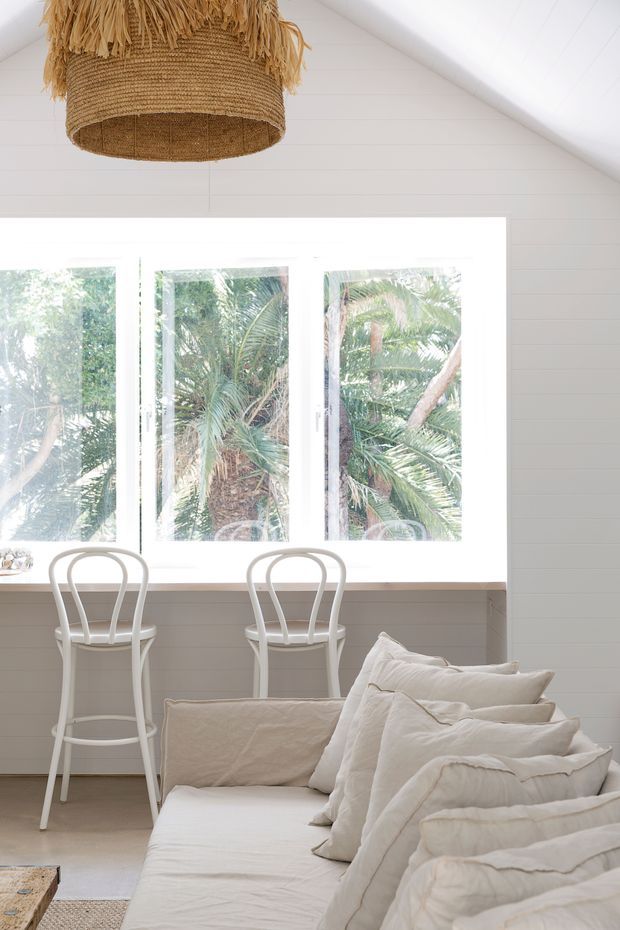 Bright, airy open-plan interior with a white-painted sloped ceiling and horizontal wall panelling. A large bank of white-framed windows overlooks lush tropical vegetation, while a narrow breakfast counter with two white stools sits beneath the glazing. In the foreground is a cream upholstered sofa or daybed layered with soft off-white cushions and throws. A woven natural-fibre pendant light hangs overhead, creating a relaxed coastal feel.