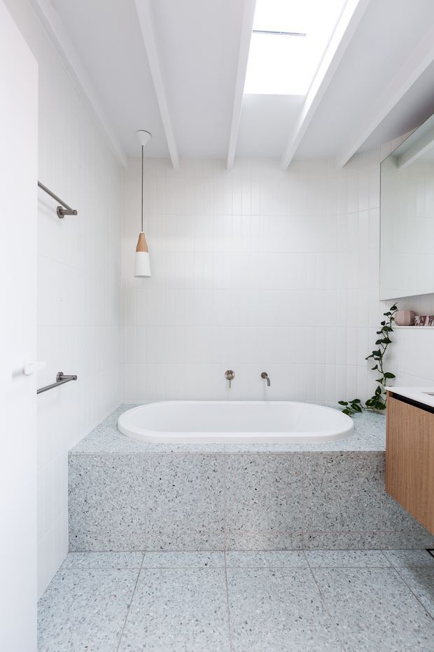 Bright contemporary bathroom featuring a built-in white bathtub set into a speckled terrazzo surround, matching terrazzo floor tiles, white wall tiles, exposed white ceiling beams, and a skylight. A timber vanity with a dark benchtop is partially visible on the right, with brushed metal fixtures and a pendant light adding subtle detail.