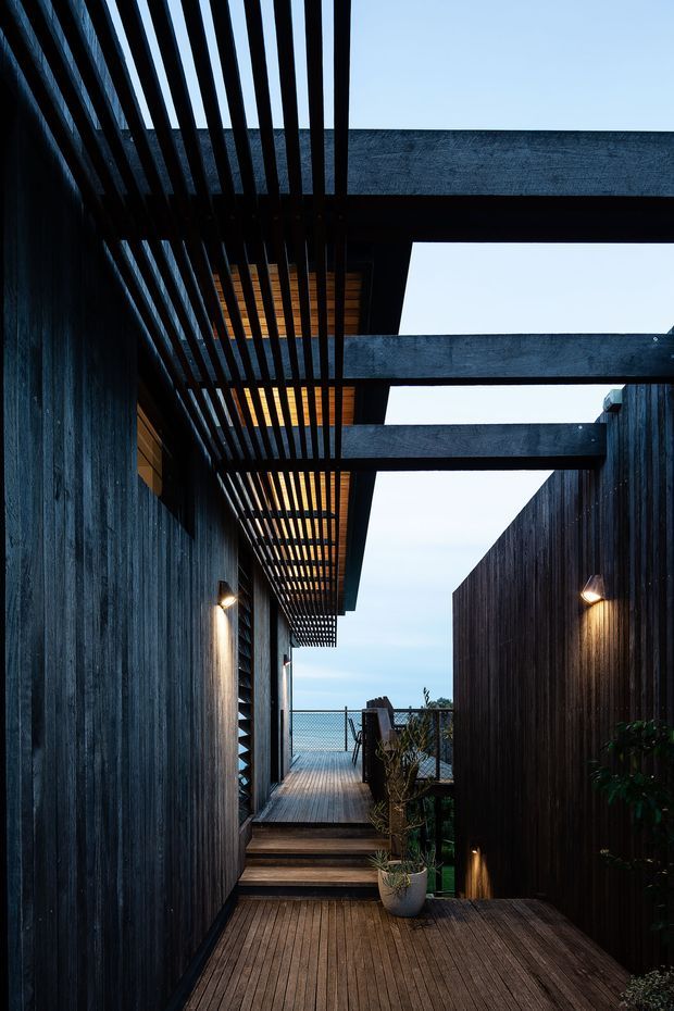 A narrow exterior walkway or courtyard passage between dark timber-clad building volumes, leading toward a fenced outlook and pale sky beyond. The path is finished with timber decking and steps, with warm wall-mounted lighting, potted plants, and a dramatic overhead pergola of closely spaced dark timber slats intersecting heavy horizontal beams. The architecture has a moody, contemporary character with strong linear geometry and a partially open roof structure.