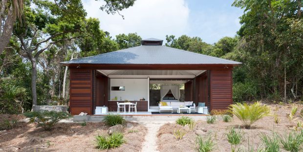 Front exterior view of a compact single-storey pavilion-style dwelling set within a wooded, sandy landscape. The structure has dark horizontal timber cladding, a broad low-pitched corrugated metal roof with a raised central roof vent, and a large open glazed living area. Outdoor chairs and a small terrace are visible through the opening, with native-style planting around a simple path.