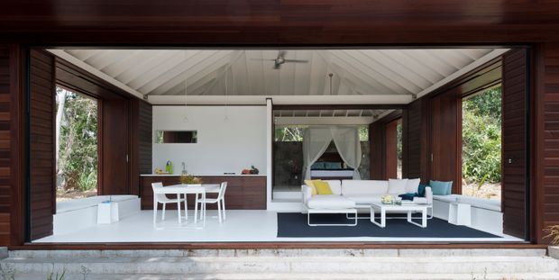 Open-plan pavilion interior viewed through fully opened dark timber-framed doors, combining a compact kitchen and dining area with a bright living room. The space has white walls and a pitched, white-lined ceiling, extensive glazing/openings to surrounding greenery, dark timber wall and door framing, built-in white seating, and contemporary white furniture. The composition emphasizes a strong indoor-outdoor connection and an airy, tropical modern feel.