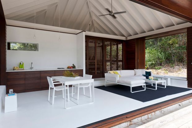 Open-plan pavilion interior with a combined kitchen, dining and living area. The space features a dramatic white vaulted ceiling with exposed beams, dark timber louvre walls and window frames, white built-in cabinetry and counters, and white contemporary dining and lounge furniture. Large open glazing connects the room to dense surrounding greenery, while a dark rug defines the seating area.