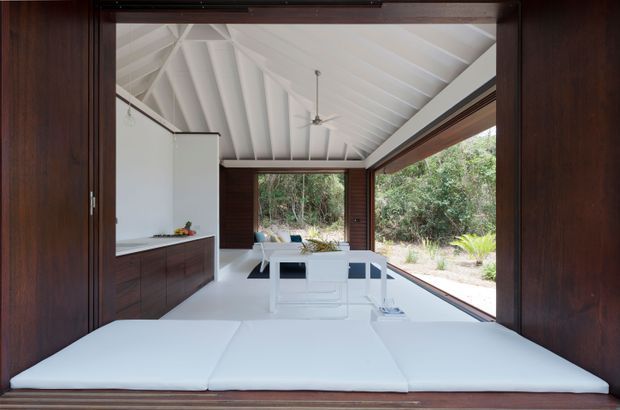 A compact, open-plan interior viewed through large dark timber-framed sliding doors. The space combines a white kitchen with dark timber cabinetry, a white dining table and chairs, and a low white built-in seating or daybed platform in the foreground. The room has a dramatic white pitched ceiling with exposed rafters and a central ceiling fan, extensive glazing, and direct views to a wooded garden.