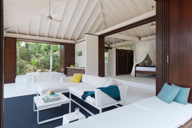 Bright open-plan tropical-style living space with a white vaulted timber ceiling, exposed rafters, dark timber wall panels and sliding doors, and expansive glazing overlooking dense greenery. White sofas and low tables create a relaxed lounge, while a bedroom with a draped four-poster bed is visible through a large opening and reflected glazing. The palette is predominantly white with dark timber accents and vivid blue and yellow cushions.