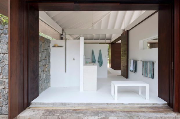 Open-plan minimalist bathroom viewed through a dark timber-framed opening, featuring white floors and walls, a freestanding white bathtub, central vanity or basin, wall-mounted towel rails, exposed white pitched ceiling structure, and a prominent rough natural-stone wall. The space has strong indoor-outdoor character with direct access to greenery and timber screening beyond.