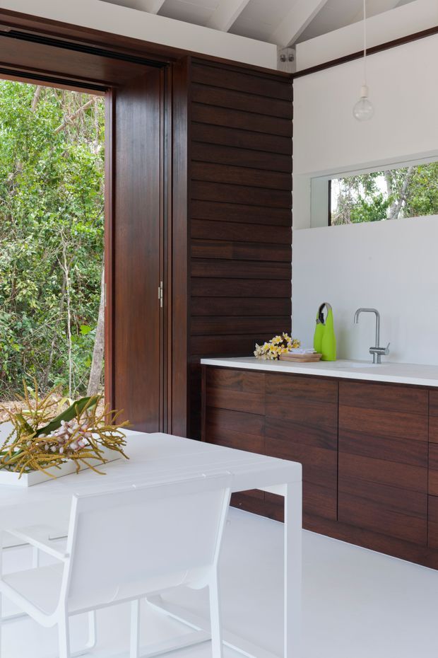 Bright, compact open-plan kitchen and dining area with dark timber cabinetry and a matching horizontal timber feature wall. White benchtops, white flooring, dining table and chairs create a crisp contrast. A large open timber-framed glazed door and horizontal window look onto dense greenery. The room has a high white raked ceiling with exposed structural beams and a simple hanging pendant.