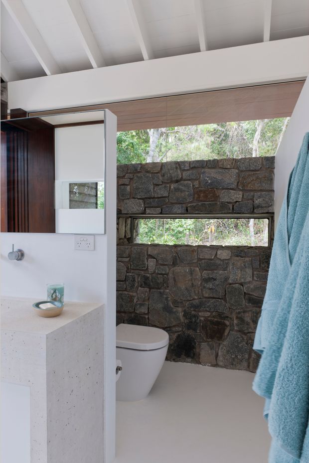 Compact contemporary bathroom with a wall-hung toilet, pale solid-looking vanity and countertop, mirrored cabinet, and exposed dark rough-stone feature wall. A narrow horizontal window brings in views of surrounding vegetation, while white exposed ceiling rafters and a timber-toned blind add warmth. The space has a clean, natural, indoor-outdoor character.