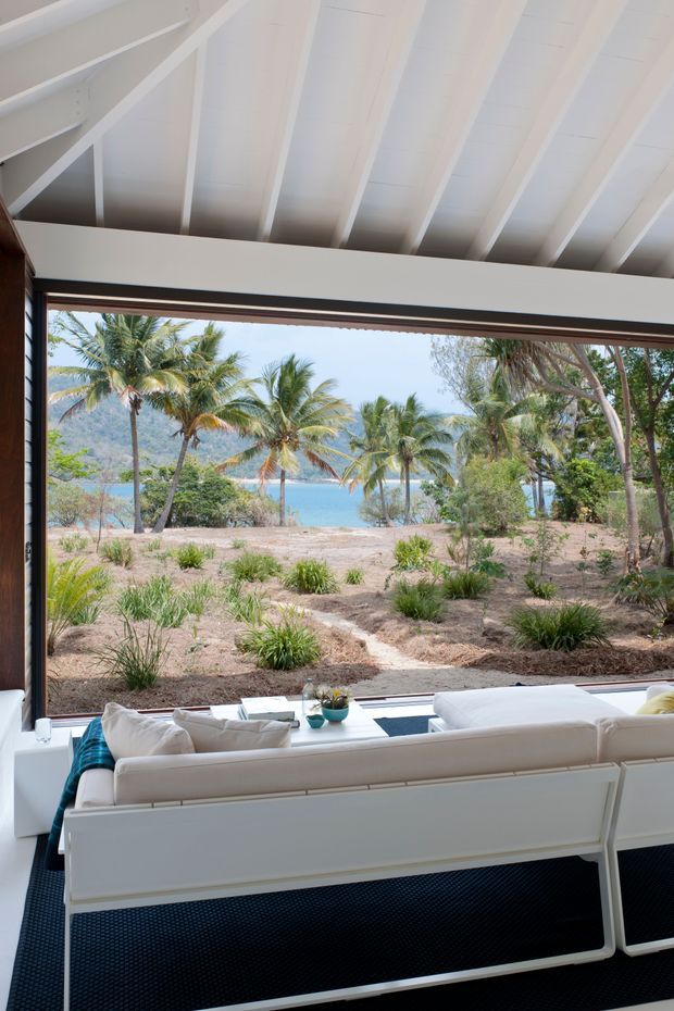 Bright coastal living room viewed from behind a white sofa, with a dramatic floor-to-ceiling window framing palm trees, sandy landscaped grounds, turquoise water, and mountainous terrain. The interior features a white pitched ceiling with exposed structural beams, dark timber window framing, white built-in or framed seating, and a dark textured floor covering. The image has a relaxed tropical resort atmosphere and strong indoor–outdoor connection.