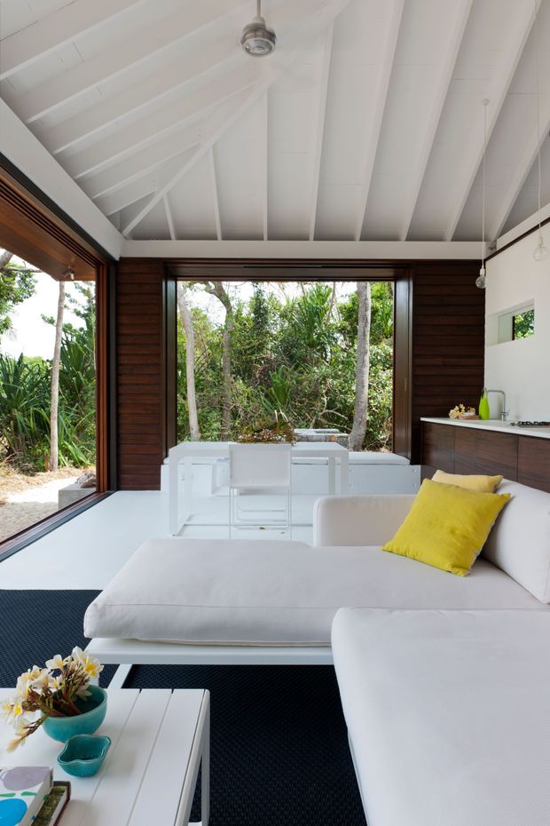Bright open-plan living and dining space with a white sectional sofa in the foreground, a white dining table and chairs beyond, and a compact kitchen along the right wall. Large open glazed openings connect the room to dense tropical greenery. The interior features dark timber wall panelling, a white pitched exposed-beam ceiling, white floors and furnishings, and a vivid yellow accent cushion.