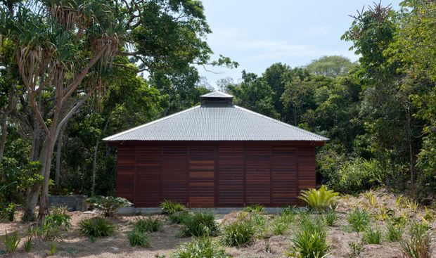 A single-storey pavilion-like building set within dense tropical vegetation. The facade is composed of dark reddish-brown horizontal timber boards and screened openings, topped by a broad light-grey corrugated hipped roof with a small raised roof vent or cupola. Sparse native-style planting and sandy ground surround the structure.