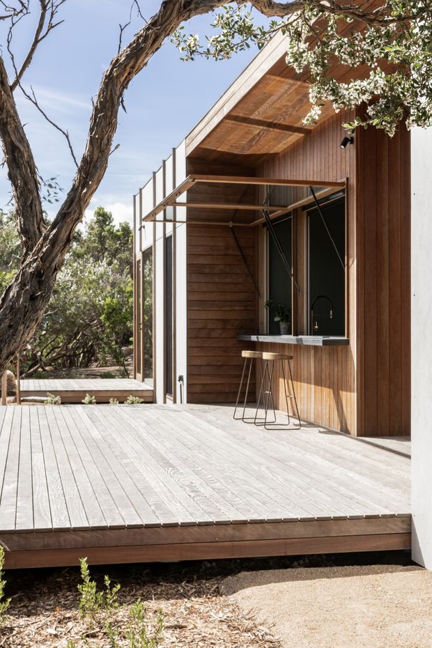 Exterior architectural photograph of a contemporary timber-clad dwelling opening onto a broad timber deck. The facade combines warm vertical and horizontal wood cladding with crisp white wall planes, dark-framed glazing, and a projecting roof overhang. A compact outdoor bar or kitchenette with a stone-like counter, black sink tap, and two stools sits beneath the covered opening. Native trees and landscaping surround the deck, creating a relaxed bush setting.