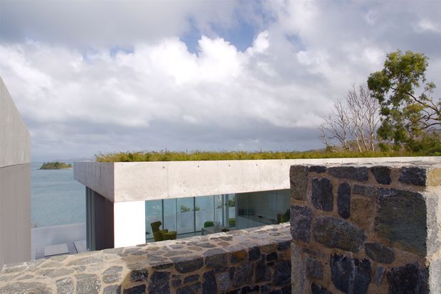 Exterior architectural view of a low, modern concrete building beside a body of water, seen from behind a rugged dark stone masonry wall. The building has a flat roof planted with low greenery, extensive floor-to-ceiling glazing, and a minimalist horizontal form. Cloudy sky, shoreline vegetation, and water are visible beyond.